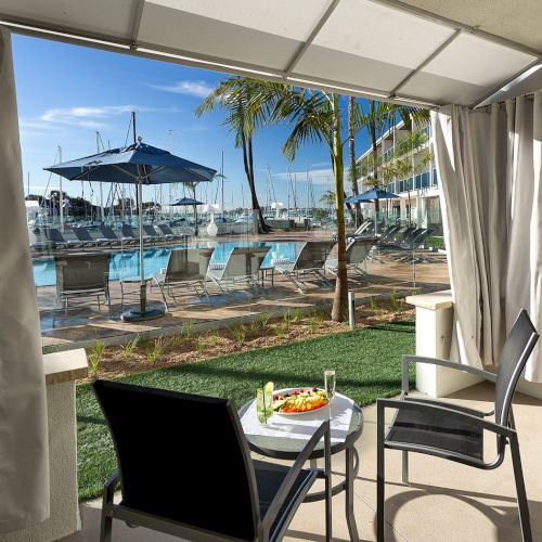 A seaside cafe with umbrellas, plants, and glass railing cozy seating overlooking blue water and a sunny view.
