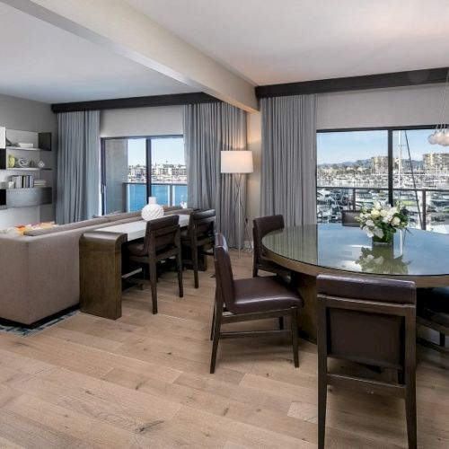 A modern living room with a cozy beige sectional, glass coffee table, and a dark dining set, large windows offering city views, bright and airy.