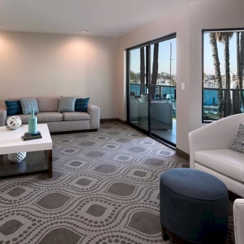A stylish living room with a patterned rug, gray sofa and armchairs, a glass coffee table, a fireplace, and large windows letting in natural light.
