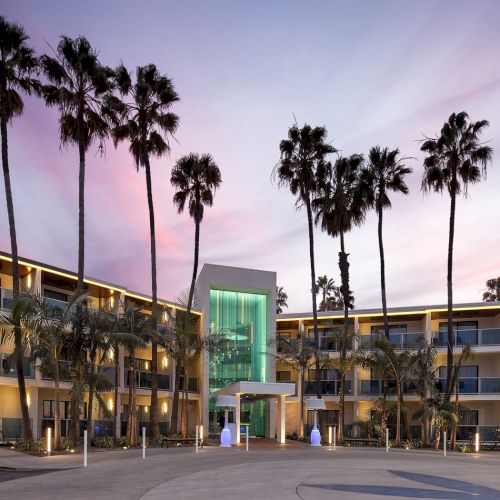 A mid-century hotel with palm trees, a turquoise glass entrance, and a pastel sunset sky, facing a wide driveway and surrounding balconies.