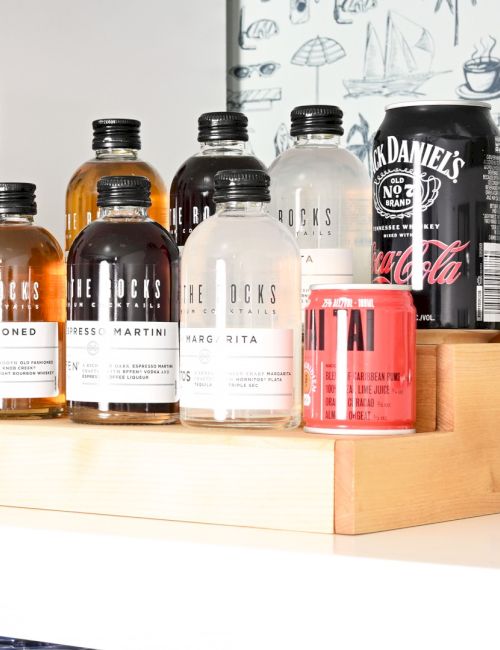 A collection of liquor bottles on a shelf, including wine, whiskey, and spirits, arranged on wooden organizers with a Coca‑Cola can in view.