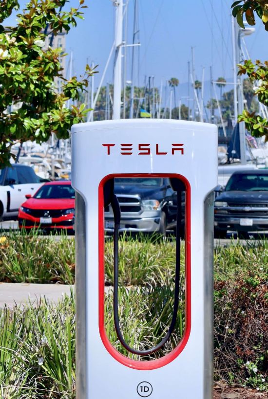 A Tesla electric car charging station at a marina/parking area with red-framed charger and two red signs on posts.