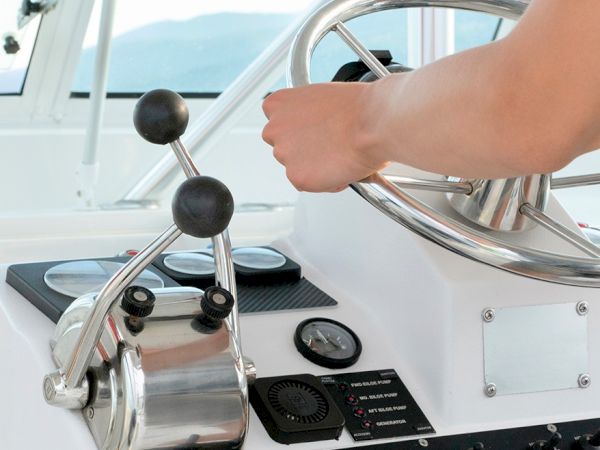 A close-up of a boat helm: steering wheel, throttle levers, gauges, and control panel being operated.