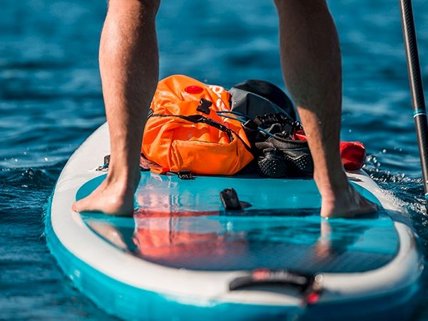 A person stands on a small paddleboard or stand-up paddleboard in blue water, with an orange life jacket and gear at the center, ready to paddle.
