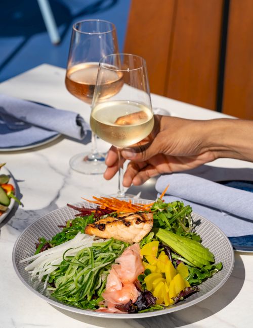 Two people toast with white wine over a fresh salad plate featuring greens, salmon, and colorful veggies.