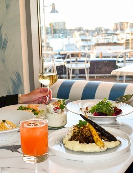 Two people dining at a bright restaurant by the window, plates of pasta and salad, and drinks clinked at the table.