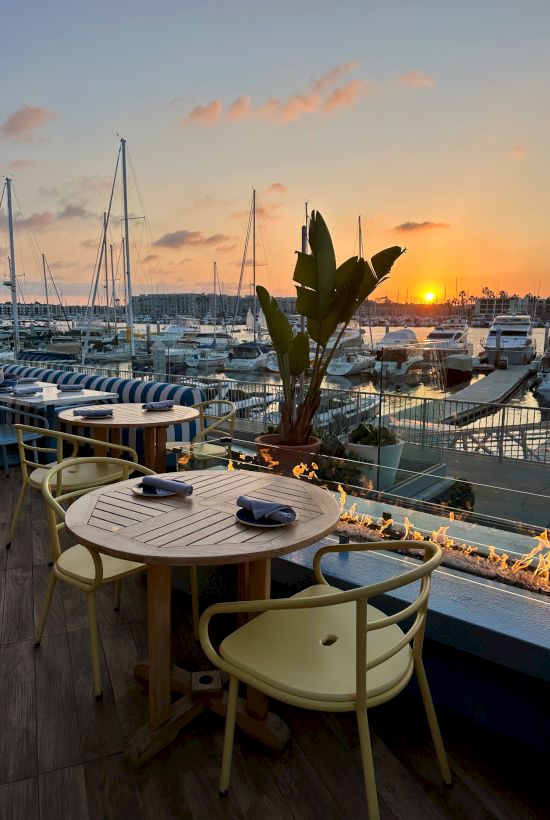 A waterfront dining area at sunset with tables, chairs, a plant, and boats in a marina; warm sky, calm water, and string lights.