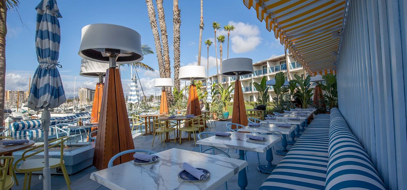 A sunny waterfront dining setup with blue-and-white striped decor, palm trees, and modern outdoor tables under shade.