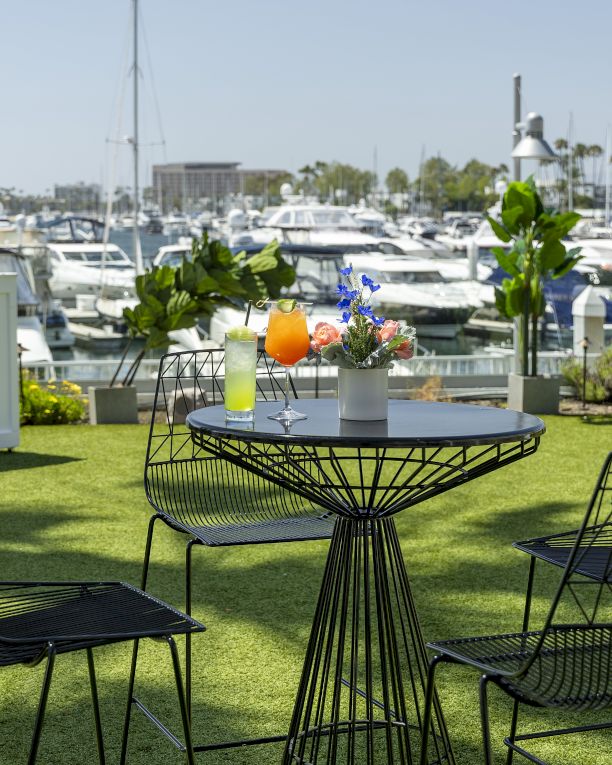 Outdoor marina cafe with black wire chairs and a round table topped by drinks and flowers, boats docked in the background by sunny water.