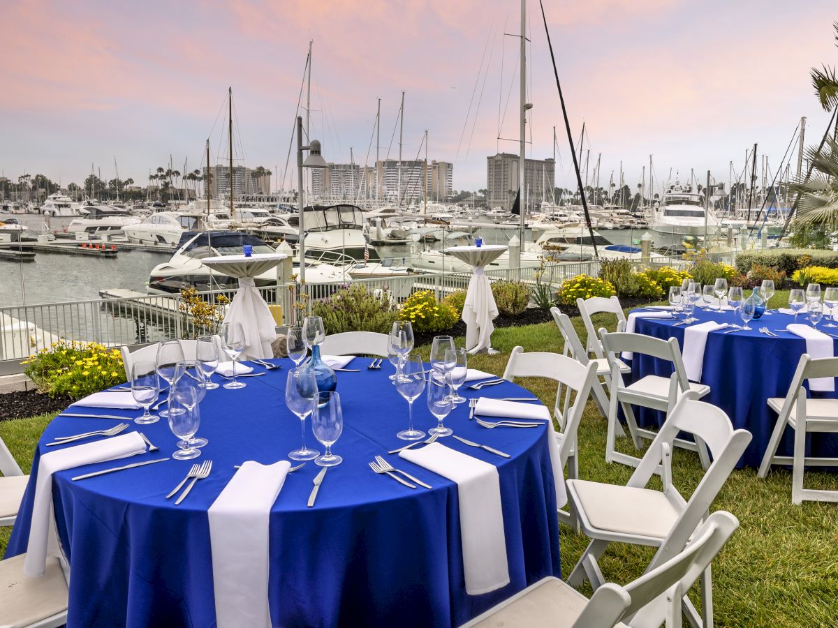 Elegant outdoor marina dining setup with round tables in royal blue linens, white napkins, glassware, and white chairs, against a harbor backdrop.