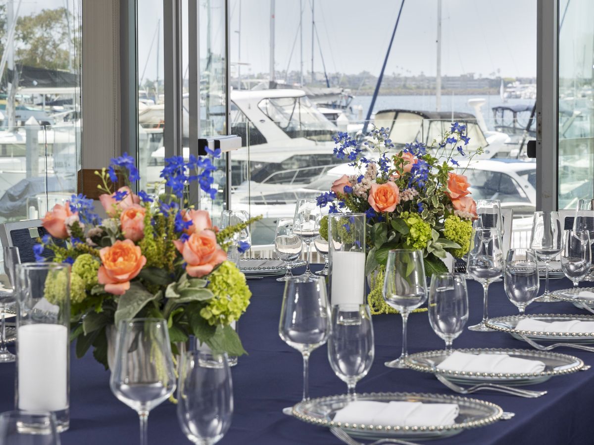 A formal dining table with navy blue cloth, elegant silverware, glassware, and colorful floral centerpieces near a marina backdrop.