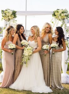 A group of four bridesmaids in matching dresses pose on a sunny beach, smiling and lifting their dresses slightly for a fun, coordinated photo.