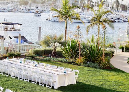 Outdoor waterfront wedding setup with a long white-table arrangement, palm trees, and a marina in the background, ready for guests.