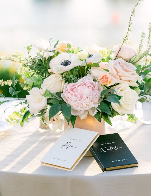 A table with a floral centerpiece of white and pink blooms, elegant place settings, and two black and gold wedding menus in front of the arrangements.