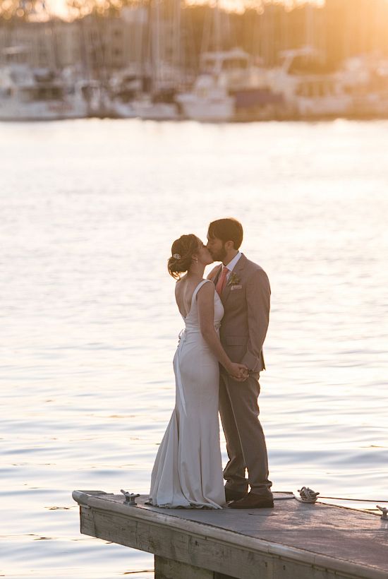 A couple in wedding attire stands on a dock by a calm harbor at sunset, sharing a quiet, intimate moment by the water.