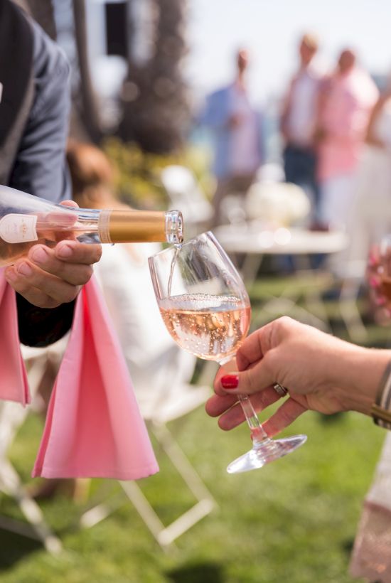 Pouring ros&eacute; into a flute at an outdoor party, hands clink champagne glasses with guests blurred in the background.
