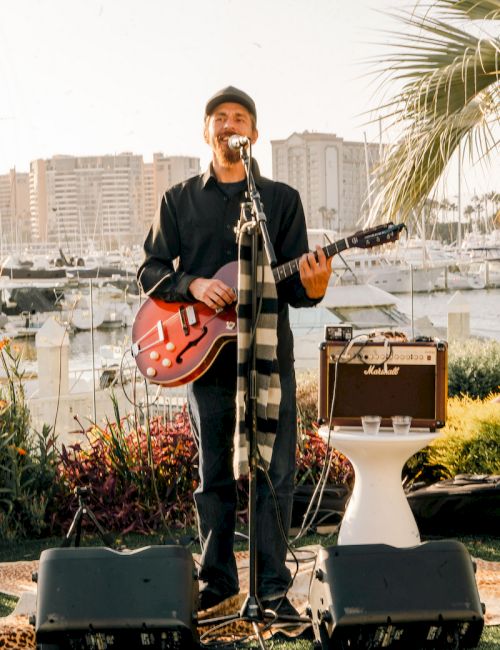 A male singer with a red guitar performs outdoors by a marina, microphone and amps set up, palm tree nearby, boats in the background.